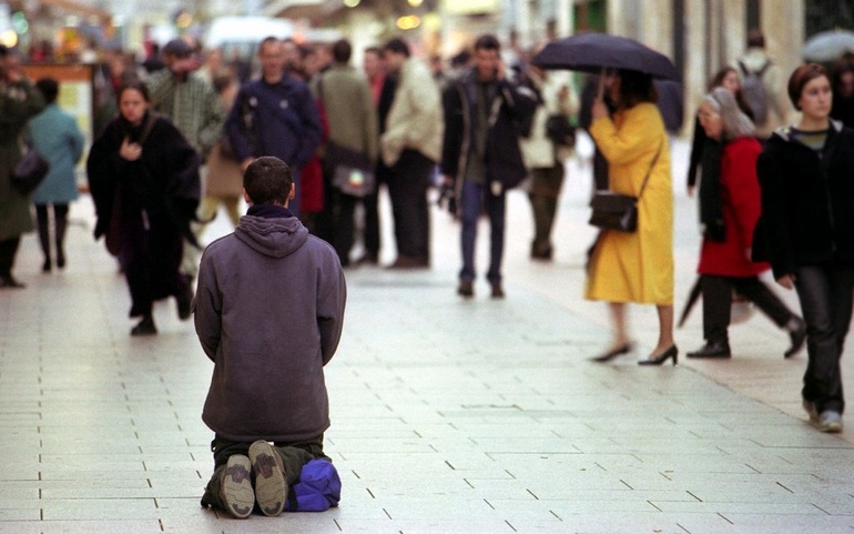 Mann med ryggen til på kne på en sovepose på gata. Folk går forbi.